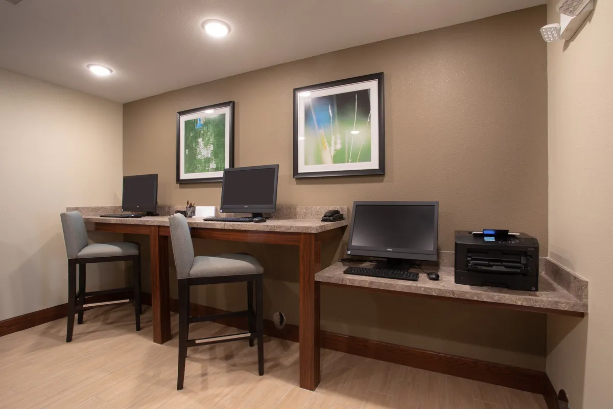 A modern business center with desks equipped with desktop computers, framed artwork on the walls, and a printer conveniently placed within reach. The lighting is professional yet warm, fostering an atmosphere ideal for productive work sessions, printing, or quick meetings.