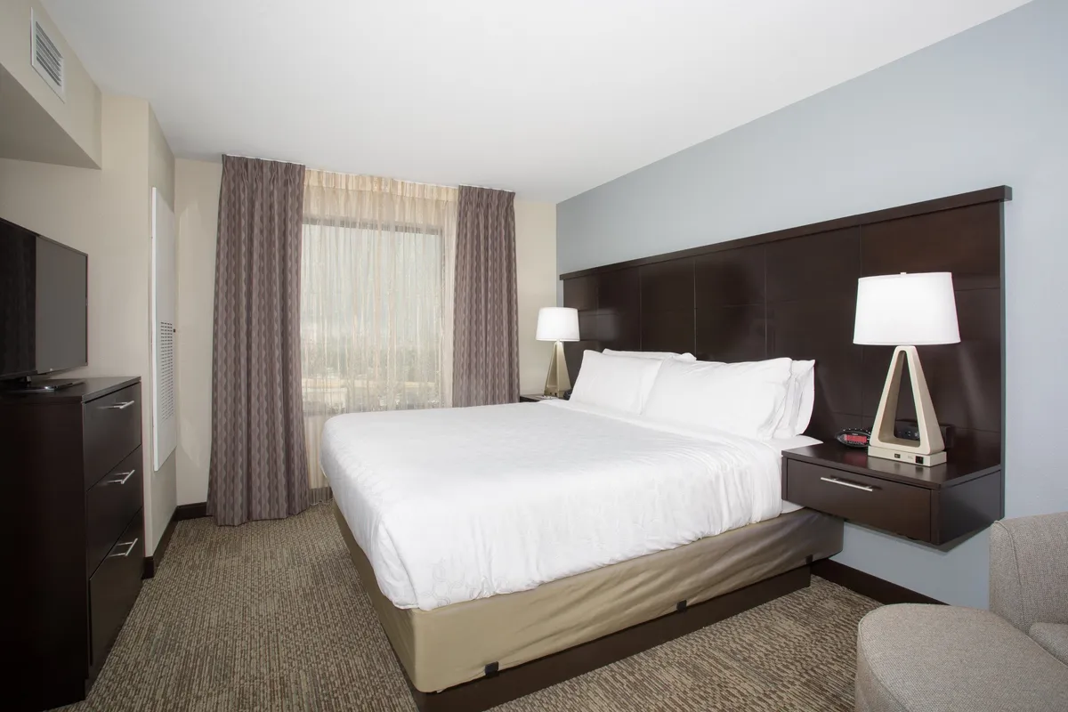 A serene king bedroom featuring a large bed with plush bedding, a window allowing abundant natural light, a nightstand with a lamp, and a flatscreen TV mounted on the wall. The space feels quiet, cozy, and inviting—ideal for relaxing or unwinding after a busy day.