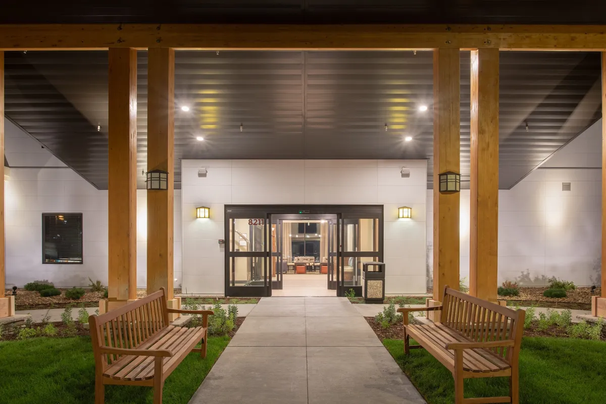 The building’s main entrance framed by neat landscaping and comfortable benches, providing a sense of security and welcome as guests arrive. The space is inviting and clearly designed to facilitate easy access and relaxation outdoors.