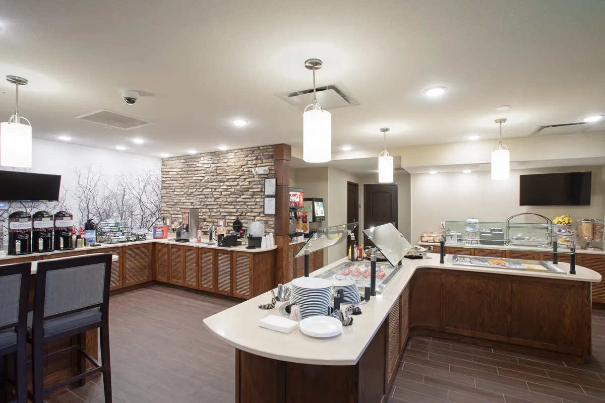 A bright and inviting breakfast buffet area with a well-stocked food display, coffee station, decorative stone wall, and comfortable seating. Guests are seen enjoying breakfast, with an overall mood of relaxation and hospitality.
