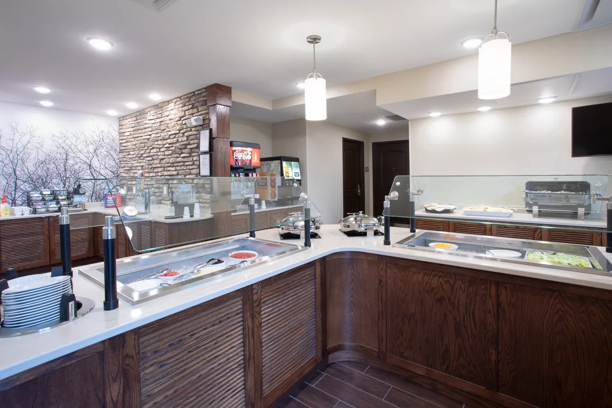 Another view of the buffet area focusing on the food display, beverage station, and stone accent wall, with guests mingling and choosing their morning meals. The mood is social and welcoming, emphasizing connection over food.
