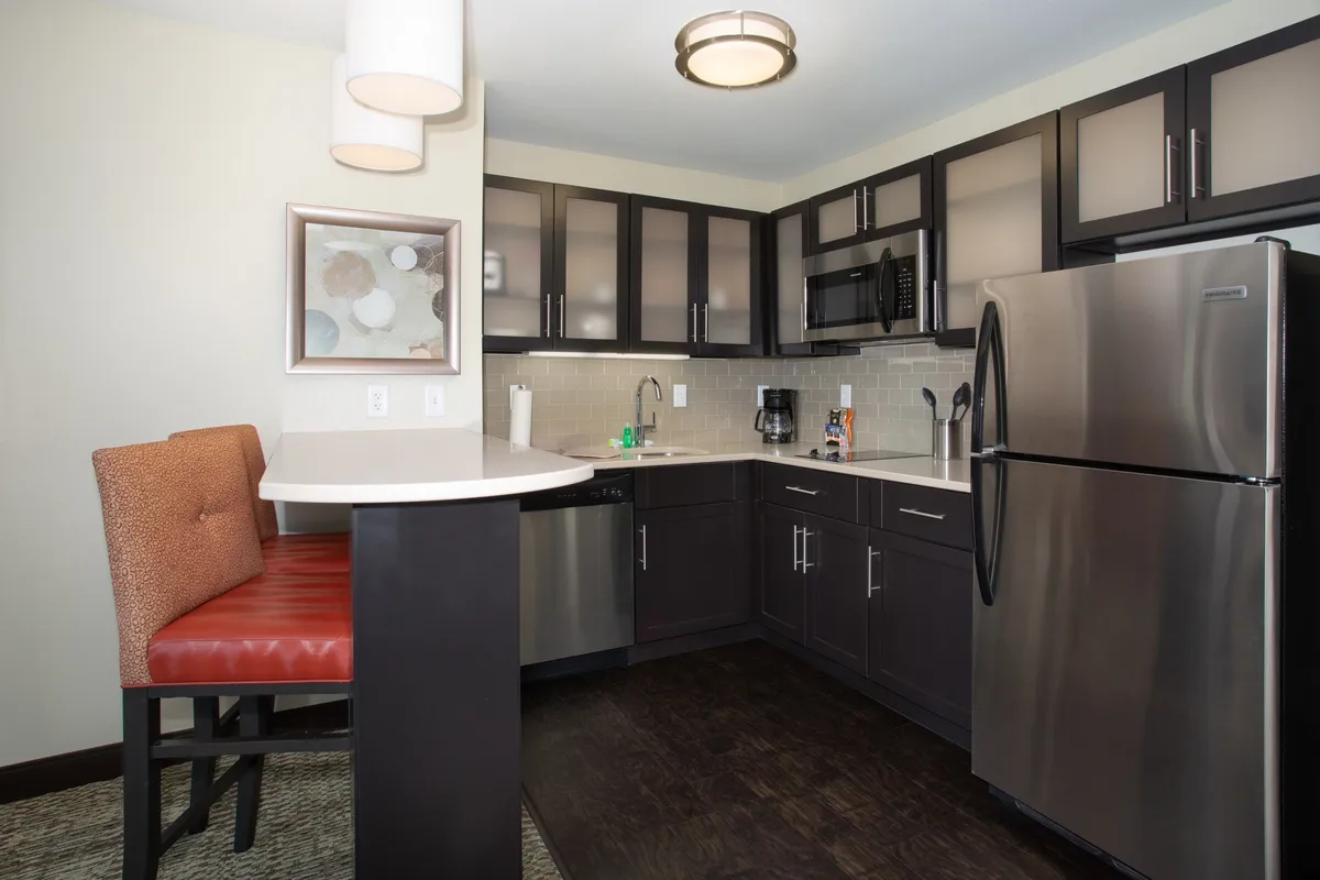 A modern, open-concept kitchen featuring a central island, stainless steel appliances including a refrigerator, stove, and microwave, and bar stools for casual dining or conversation. The scene suggests a space ideal for cooking, entertaining, and sharing meals.
