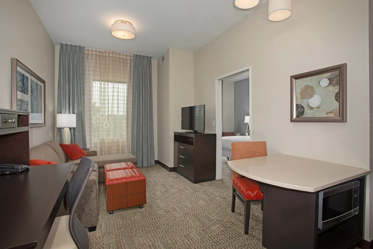 The hero image showcases a bright, inviting living area with a comfortable sofa, modern table, and television, all illuminated by natural light streaming through a large window. The bedroom doorway is visible, creating a seamless flow between spaces and suggesting both relaxation and convenience. The clean, cozy decor sets a tranquil scene for unwinding or enjoying a quiet meal.